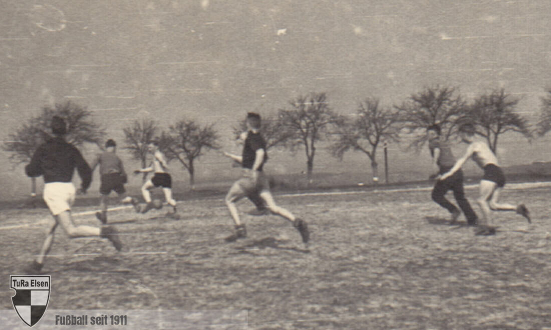 Schwarz-weiß-Foto von sechs jungen Männern beim Fußballtraining auf Merschs Wiese, sie laufen und spielen auf einem offenen Feld mit Bäumen im Hintergrund.
