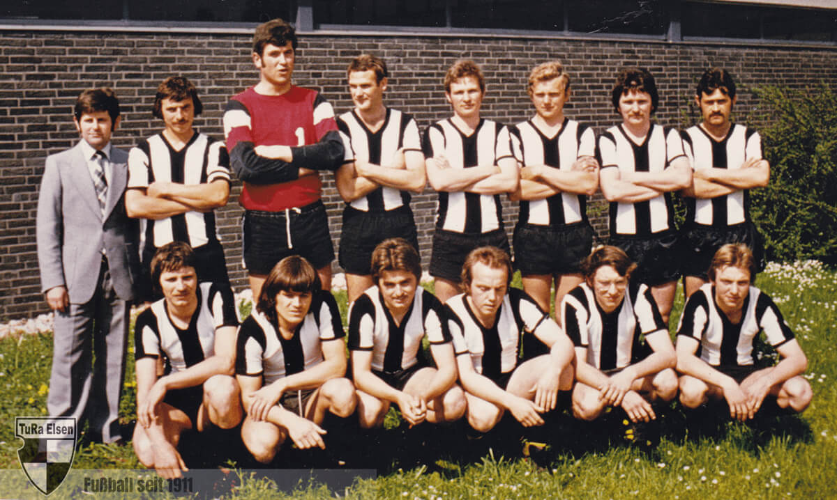 Fußballmannschaft TuRa Elsen 1976 in schwarz-weißen Trikots und schwarzen Hosen, mit Trainer im grauen Anzug vor Backsteinmauer