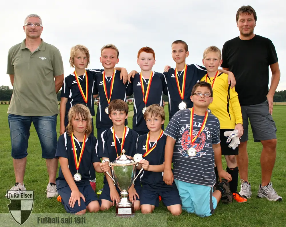 E-Junioren mit zwei Trainern, Jungen in blauen Trikots und Medaillen, ein großer Pokal vorne auf dem Rasen