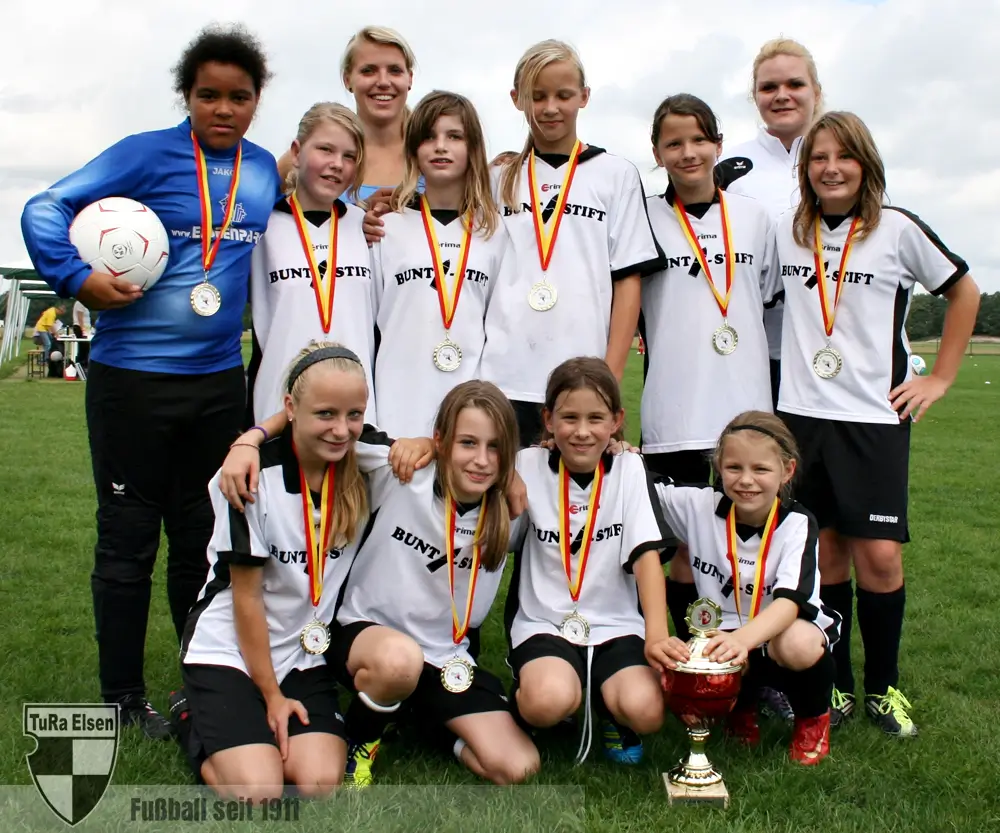 U13-Juniorinnen Fußballteam mit Medaillen und Pokal auf dem Rasen, zwei Trainerinnen im Hintergrund