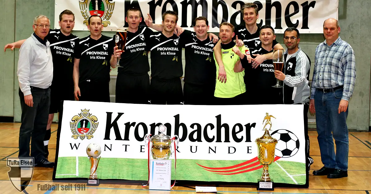 Ü32 Fußballmannschaft posiert in Sporthalle mit drei Pokalen vor Banner 'Krombacher Winterrunde'