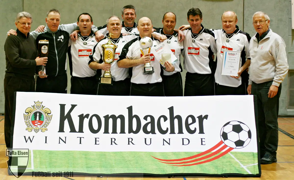 Ü50 Fußballmannschaft posiert mit Pokalen und Urkunde hinter Banner 'Krombacher Winterrunde' in Sporthalle
