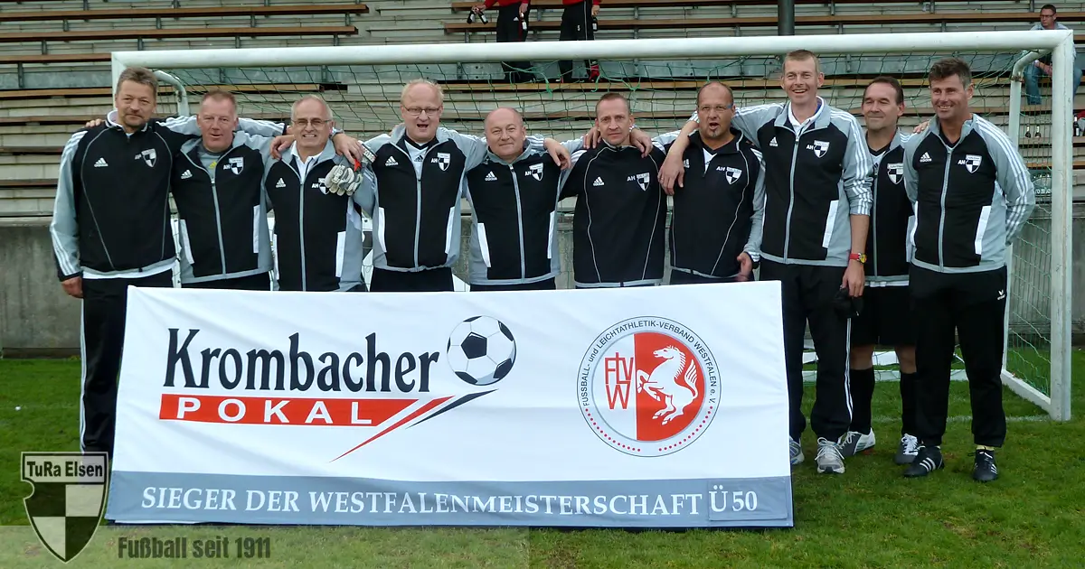 10 Spieler in schwarzen Trainingsjacken posieren hinter einem Banner mit der Aufschrift 'Krombacher Pokal Sieger der Westfalenmeisterschaft Ü50' auf einem Fußballfeld vor einem Tor