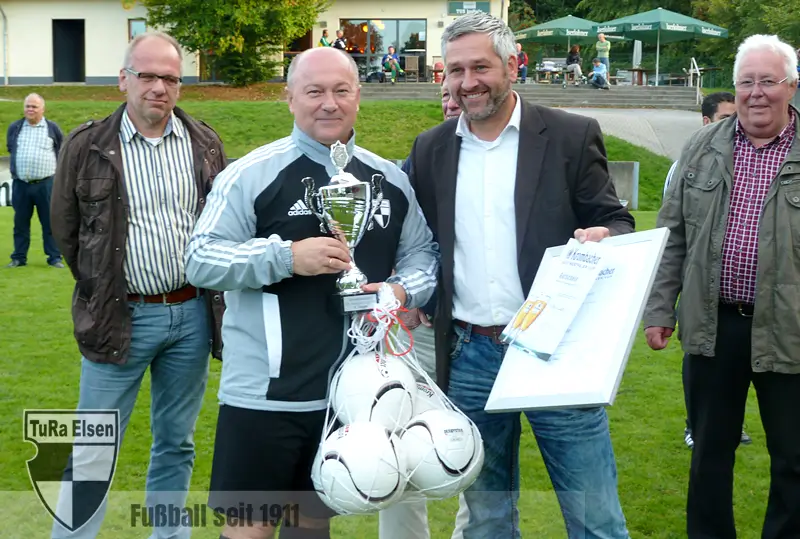 TuRa Kapitän Raimund Krick in Sportjacke hält Pokal und mehrere Fußbälle, neben ihm steht der Vertreter des FLVW mit Urkunde, weitere Männer im Hintergrund auf Fußballrasen