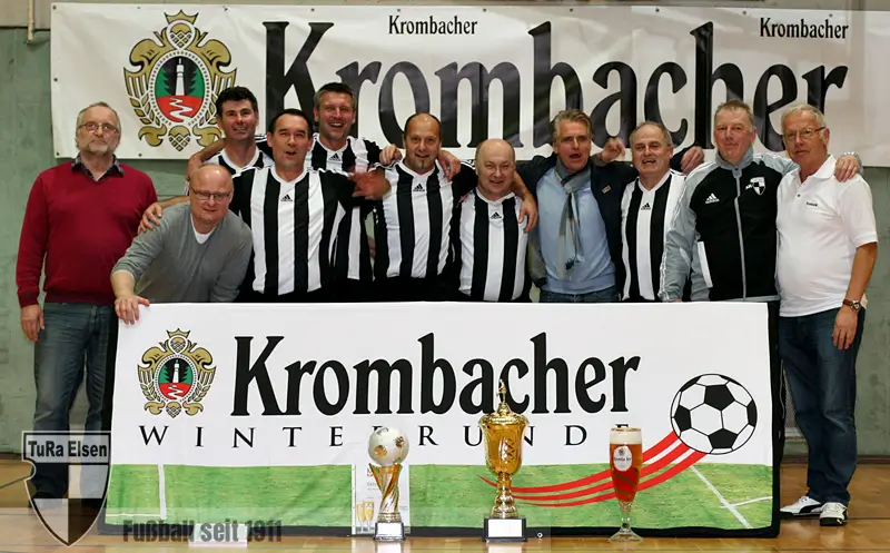 Elf Männer, einige in schwarz-weiß gestreiften Fußballtrikots, posieren hinter einem Banner mit der Aufschrift 'Krombacher Winterrunde' und mehreren Pokalen