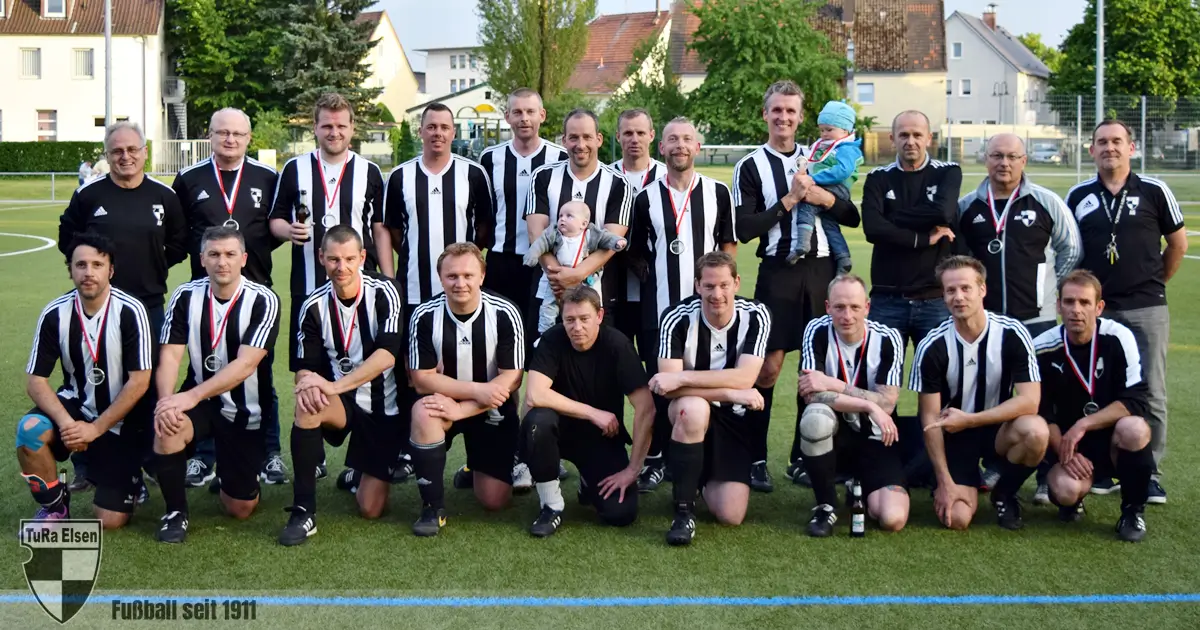Ü32 TuRa Elsen mit 16 Spielern und Betreuern in schwarz-weißen Trikots auf dem Spielfeld, einige tragen Medaillen, zwei halten Babys