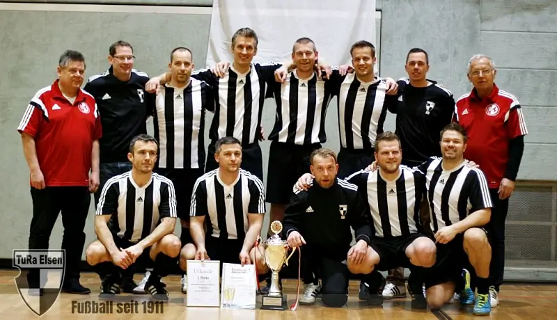 Ü32 TuRa Elsen in schwarz-weiß gestreiften Trikots mit Pokal und Urkunden in einer Halle, zwei FLVW Vertreter in roten Shirts