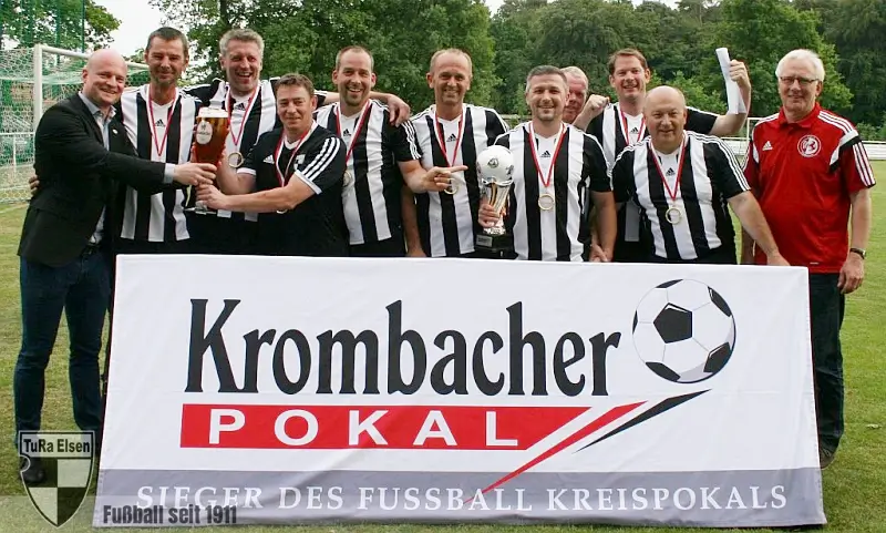 TuRa Altehrren Ü40 Ü40 Fußballteam in schwarz-weißen Trikots hält Pokale und Medaillen hinter Banner mit Aufschrift 'Krombacher Pokal Sieger des Fussball Kreispokals' auf grünem Spielfeld