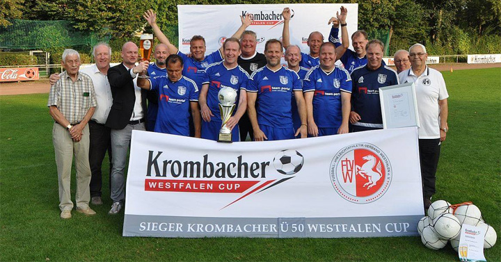Die Oldies der SG Balve/Garbeck in blauen Trikots halten Pokal und Banner mit Aufschrift 'Krombacher Westfalen Cup Sieger Krombacher Ü50 Westfalen Cup' auf Fußballfeld