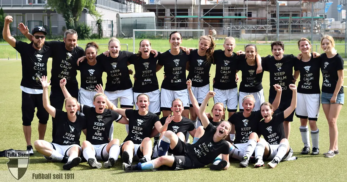 Frauen-Fußballteam der TuRa Elsen feiert Wiederaufstieg auf dem Spielfeld, tragen schwarze T-Shirts mit 'KEEP CALM WE ARE BACK!'