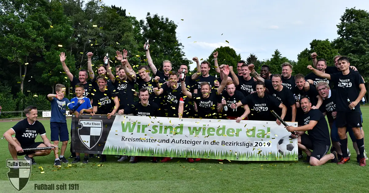 1. Herren TuRa Elsen feiern Aufstieg in die Bezirksliga 2019 mit Banner 'Wir sind wieder da!' auf grünem Spielfeld