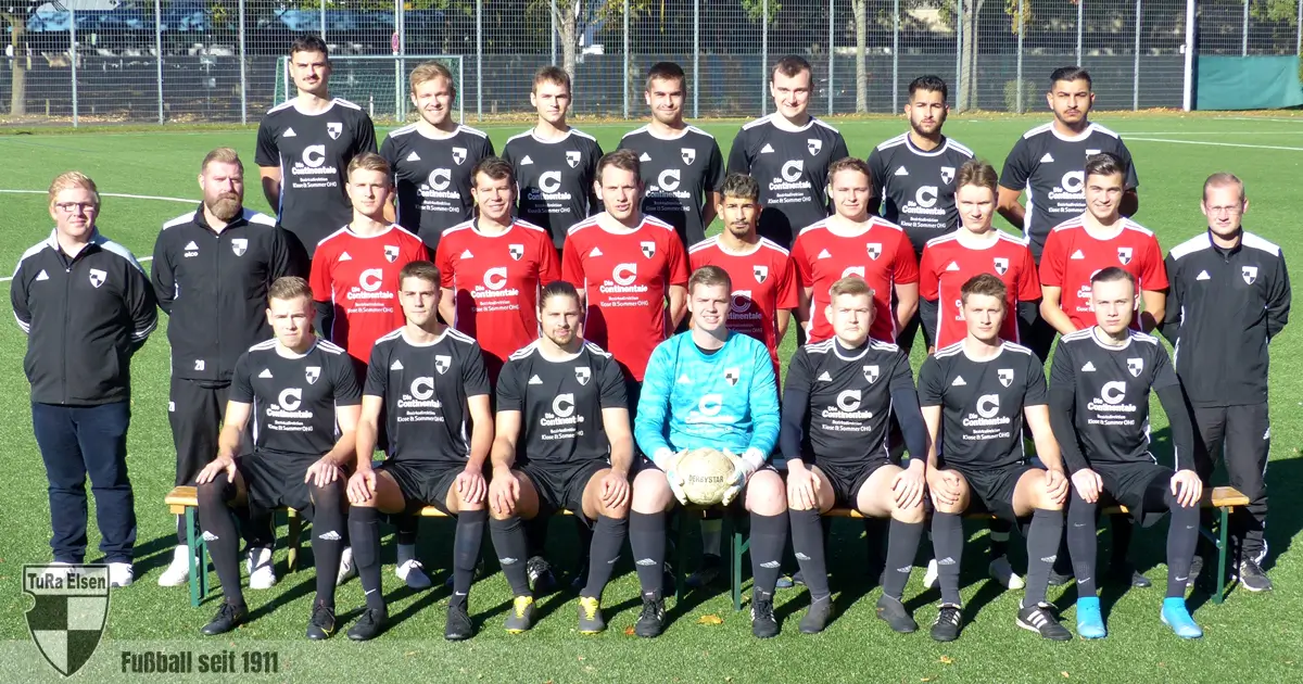 Mannschaftsfoto der 1. Herren TuRa Elsen in Fußballtrikots auf dem Spielfeld, einige Spieler in schwarzen, andere in roten Trikots, Torwart in hellblauem Trikot
