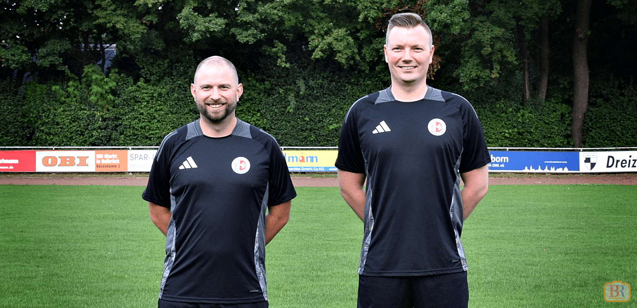 Die Schiedrichter Olli und Kevin in schwarzen Adidas-Trikots stehen nebeneinander auf einem Fußballrasen vor Werbebanden und Bäumen im Hintergrund