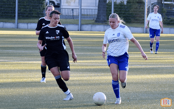 Zwei Spielerinnen in Bewegung auf dem Spielfeld, eine in weißem und eine in schwarzem Trikot, Ball vor der Spielerin in Weiß