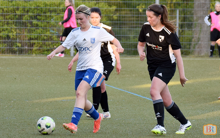 Zwei Spielerinnen in Aktion auf dem Spielfeld, eine in weißem Trikot mit Ball, die andere in schwarzem Trikot verfolgt sie.