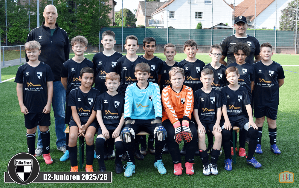 Gruppe von 16 Fußballspielern in Trikots, stehen und sitzen auf einem Fußballfeld. Zwei Trainer stehen hinter ihnen.