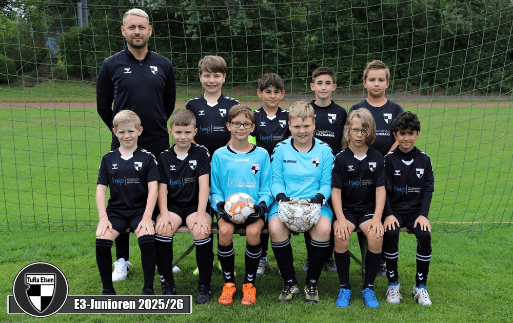 Eine Gruppe von acht Fußballspielern in schwarzen Trikots und zwei Torwarte in blauer Ausrüstung auf einem Rasenfeld. Trainer steht daneben.