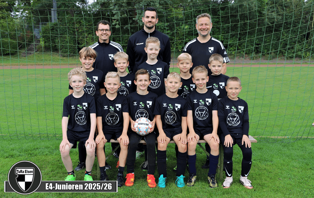 Gruppe von 11 Fußballspielern in schwarzen Trikots, auf einem Fußballfeld vor einem Tor aufgestellt. Drei Trainer stehen hinter ihnen.
