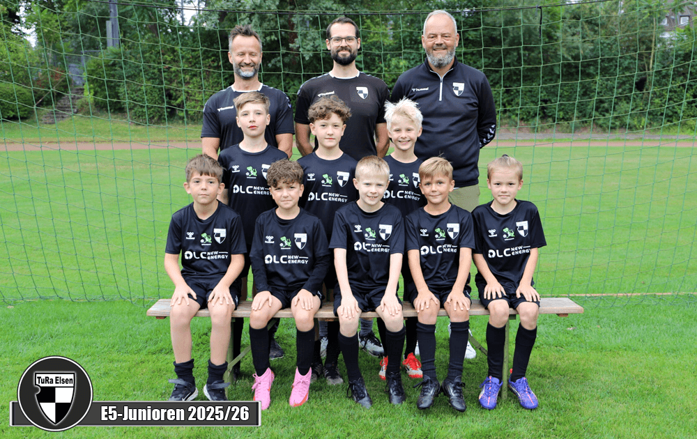 Eine Gruppe von acht Kindern in schwarzen Fußballtrikots, sitzend und stehend auf einer Bank vor einem Fußballfeld. Drei Trainer stehen hinter ihnen.