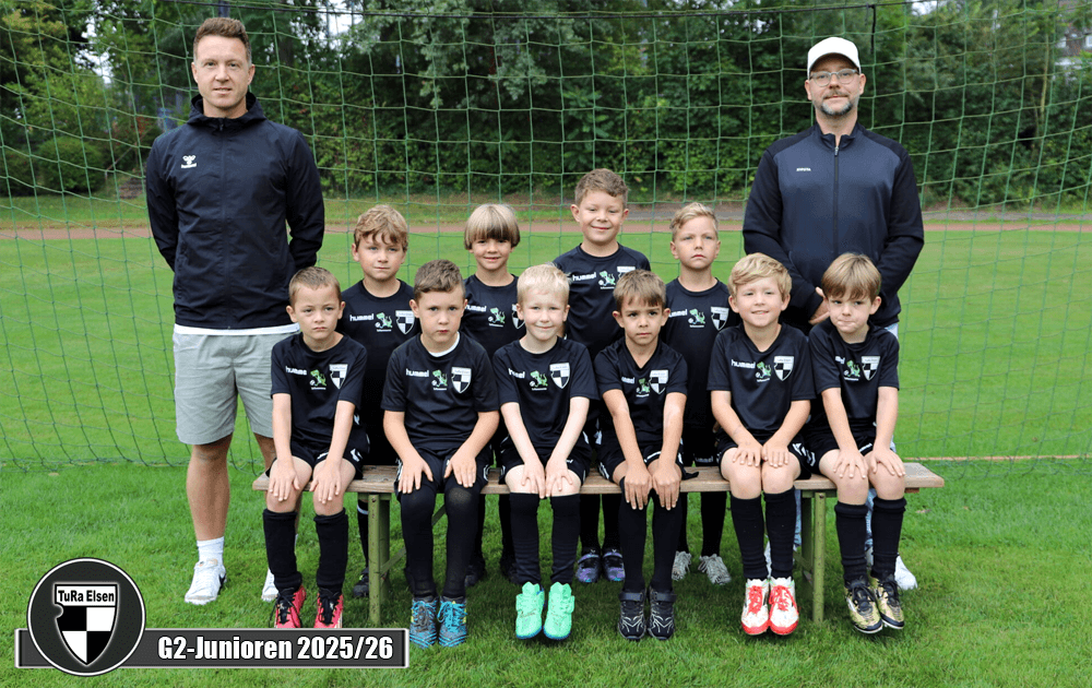 Zwei Trainer stehen links und rechts von einer Gruppe von zehn Kindern in schwarzen Fußballtrikots. Die Kinder sitzen auf einer Bank, alle tragen schwarze Shorts.
